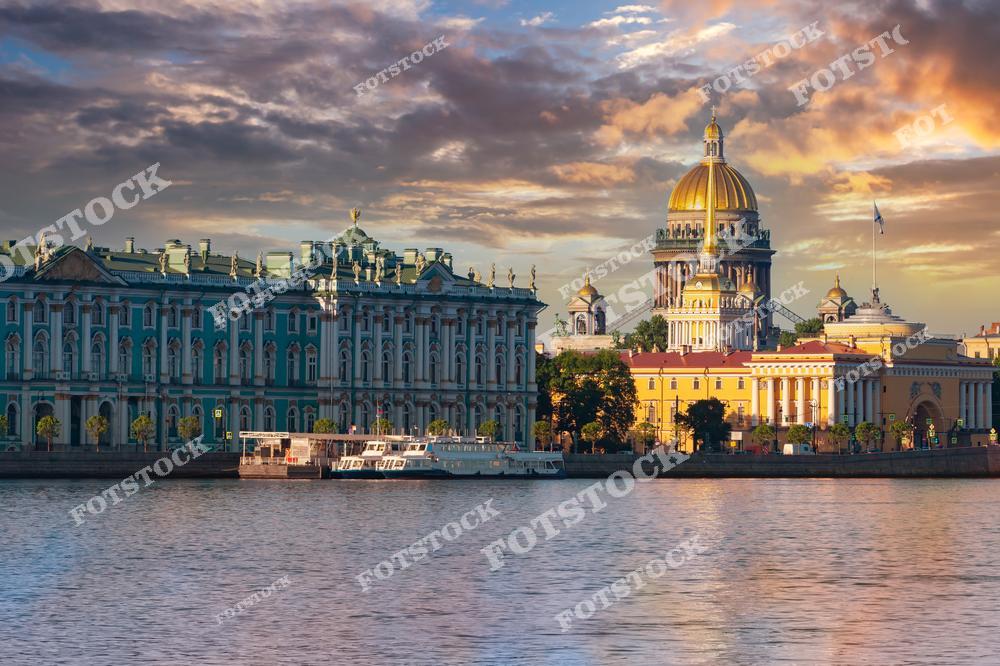 На переднем плане — река Нева и причал с прогулочными катерами. Слева — величественный Зимний дворец, входящий в состав комплекса Эрмитажа. Справа возвышается купол Исаакиевского собора — одного из крупнейших храмов России. Также на фото виден фасад здания Адмиралтейства. Закат окрашивает небо и архитектуру в золотистые оттенки, подчёркивая красоту центра Петербурга.